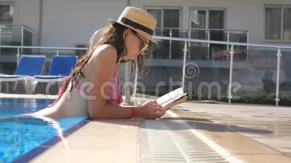 漂亮的女孩戴着帽子和太阳镜在游泳池看书年轻女子在假期里在温暖的阳光下放松视频的预览图