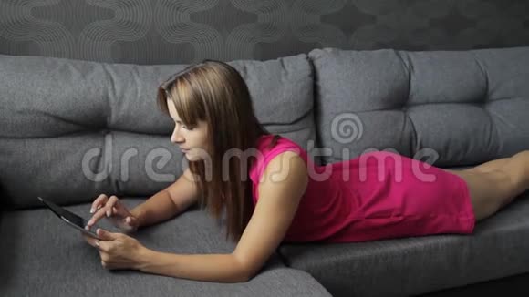 穿裙子的女人躺在沙发上的平板电脑上趴在肚子上可爱的女人在家视频的预览图
