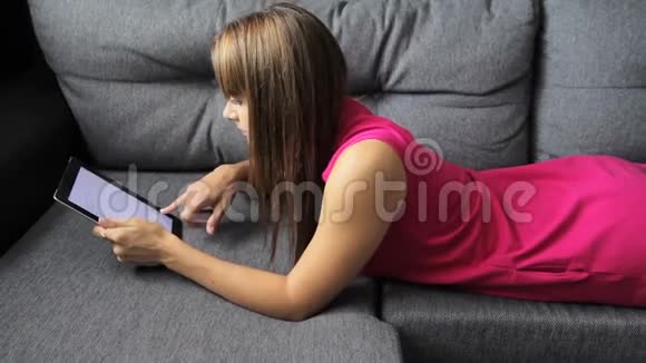 穿裙子的女人躺在沙发上的平板电脑上趴在肚子上可爱的女人在家视频的预览图