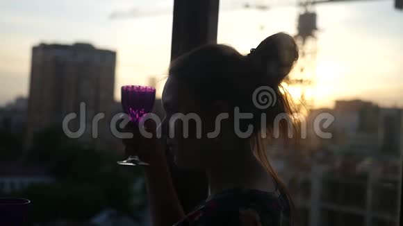 女孩坐在窗边手里拿着一个玻璃杯欣赏着城市里的日落慢动作视频的预览图