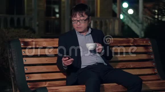 夜晚公园的亚洲商人使用智能手机喝一杯咖啡视频的预览图
