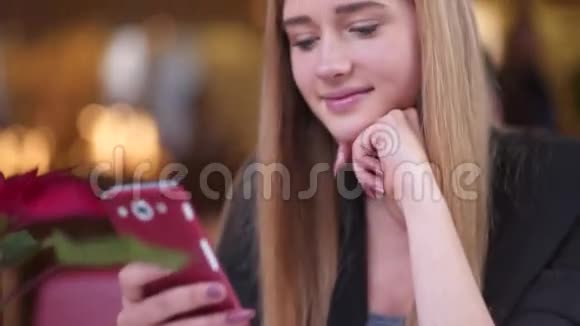 穿着黑色夹克的漂亮金发女郎坐在咖啡馆A商店里她专注于和她的朋友发短信视频的预览图