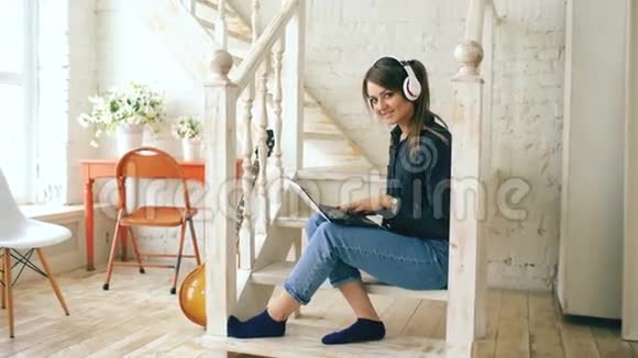 一位戴着耳机和笔记本电脑的美女坐在家里的楼梯上摆着姿势和微笑视频的预览图