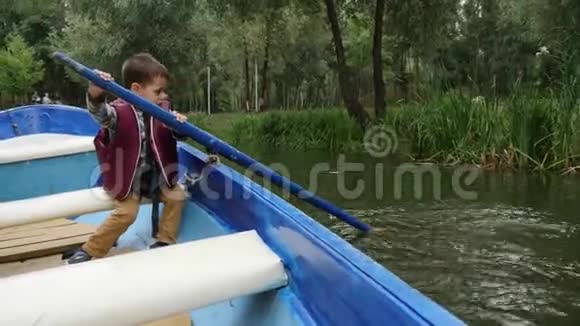 秋天迷人的可爱男孩坐在湖上的船上可爱的小孩子在公园的湖边的木船上航行绿树成荫视频的预览图