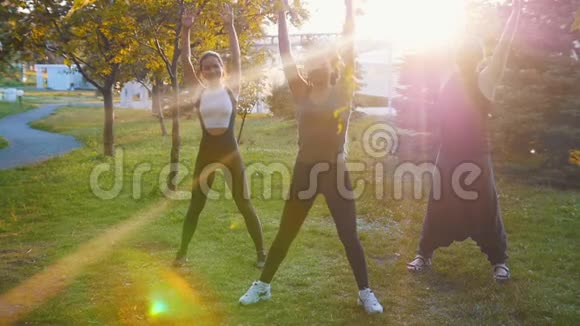 两位年轻女性在阳光下和教练一起在公园里做瑜伽练习一位女士有着长长的蓝色恐惧症视频的预览图