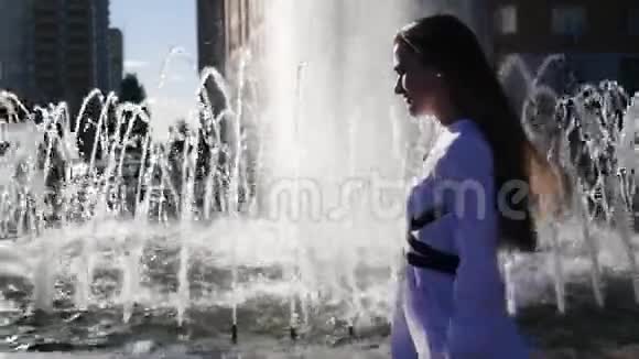 年轻的红发女孩模特穿着一件白色衬衫在一个清新的喷泉附近走着喷泉里反射着阳光和阳光视频的预览图