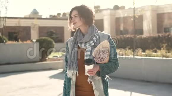 年轻女子在城市建筑背景下手持一束鲜花和一杯咖啡在镜头前摆姿势视频的预览图
