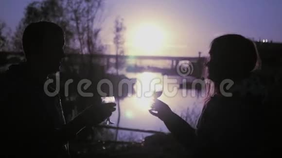 年轻的恋人手里拿着酒杯和酒手里拿着酒杯在河岸上站着背景是视频的预览图