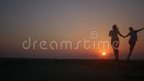 两个朋友牵着手在橙色的夕阳下在海滩上跳跃高清1920x1080慢动作视频的预览图