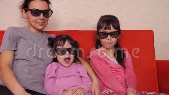 母亲带着两个女儿在沙发上用3D眼镜看电影家人在橙色沙发上看3D电影视频的预览图