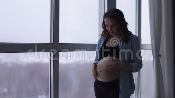 一个快乐的孕妇站在一个大的全景窗口位于一个高空他摸他的肚子视频的预览图