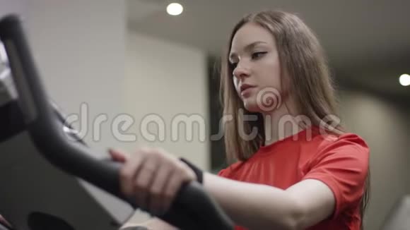 穿着红色衬衫和蓝色腿的漂亮女孩骑着运动自行车在新的健身房里看时间视频的预览图