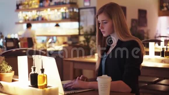 穿着黑色夹克的漂亮金发女郎坐在咖啡馆A里她把注意力集中在笔记本上视频的预览图