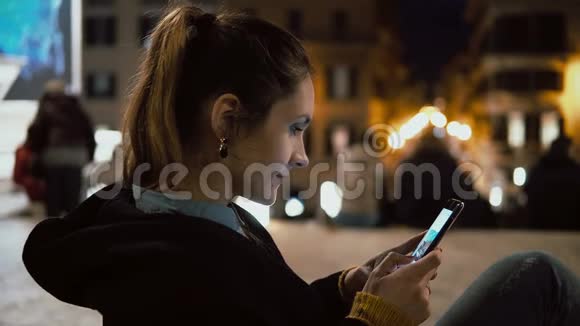 漂亮的黑发女人晚上坐在市中心用触摸屏技术在智能手机上看照片视频的预览图