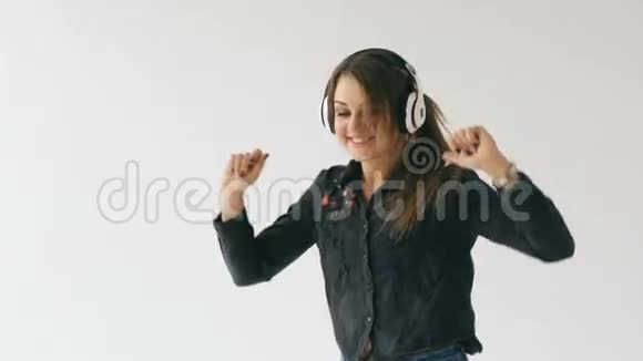 在室内听音乐和在白色背景下跳舞的年轻女子戴耳机慢动作视频的预览图
