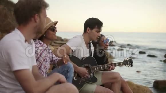 一群年轻的时髦朋友坐在海边的岩石上弹吉他唱歌慢动作视频的预览图