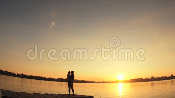 剪影日落时分一个小孩和一个女人站在水边的码头上视频的预览图