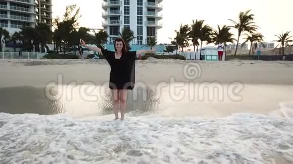 穿着黑色衣服的女人站在波浪状的海水中在阳光明媚的艾尔斯海滩上迈阿密度假胜地附近送空气亲吻视频的预览图