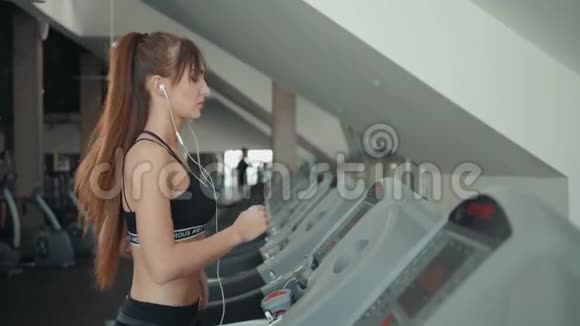 年轻的黑发女子运动员智能手机在健身房跑步机上运行女子听手机音乐视频的预览图