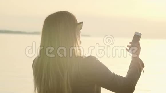 靠近一个美丽的女孩坐在海边的码头上在日落时拍了一张InstagramStorry的自拍视频的预览图
