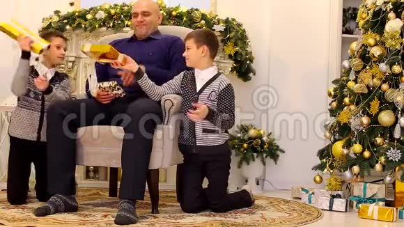 一家之主男人和儿子在有壁炉和高大圣诞树的客厅里互相赠送礼物和微笑视频的预览图