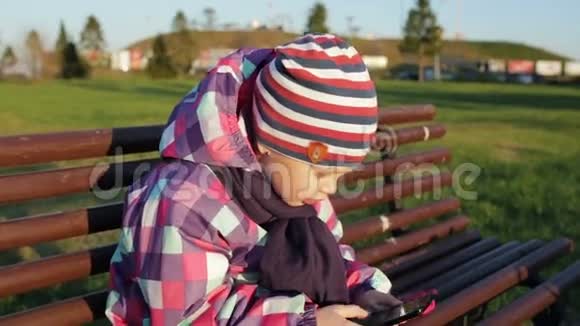 小女孩拿着智能手机坐在公园的长凳上快关门视频的预览图