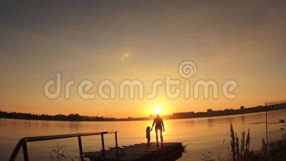 剪影日落时分一个小孩和一个女人站在水边的码头上视频的预览图