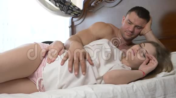 一位年轻漂亮的孕妇和她肌肉发达的丈夫躺在床上丈夫正在熨他妻子的腹部视频的预览图