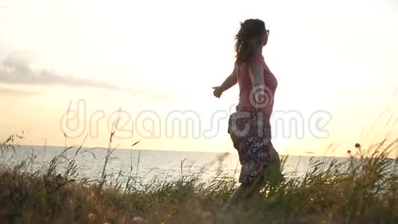 一个年轻的女孩正在草地上在夕阳和大海的背景下旋转享受着阳光1920x1080视频的预览图