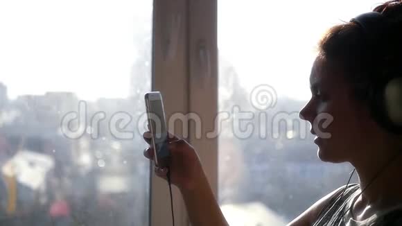 一个年轻的女孩站在窗前戴着耳机手里拿着一部手机看着这个小玩意慢动作视频的预览图