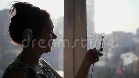 一个年轻漂亮的女孩站在她耳机的窗户附近翻过电话播放列表慢动作视频的预览图