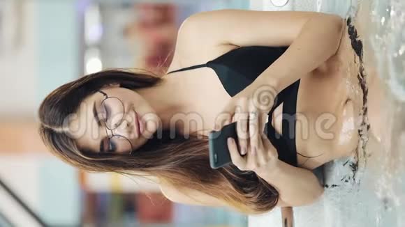 垂直视频美丽的年轻女子使用智能手机放松按摩浴缸带手机的女孩在游泳池里放松视频的预览图