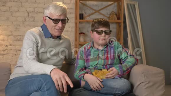 祖父和孙子坐在沙发上用3D眼镜看3D电影吃薯片看电视看节目家庭视频的预览图