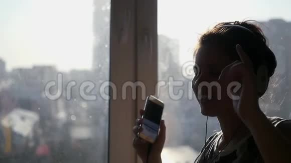 一个年轻开朗的女孩从电话里用耳机听音乐在阳光下在窗户边大声唱歌跳舞视频的预览图