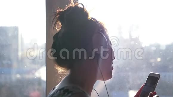 一个戴着耳机手里拿着手机的年轻活泼的女孩站在窗前跳舞唱歌视频的预览图