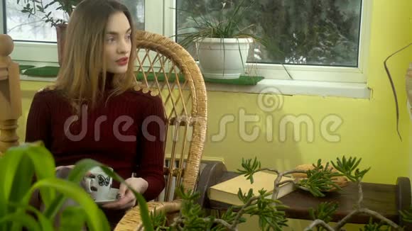 迷人的年轻女孩化妆漂亮坐在家里的柳条椅上看着窗外喝着热味的饮料视频的预览图