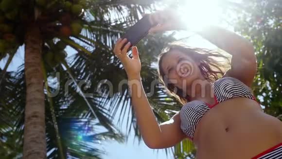 美丽的苗条女人长着深色长发站在棕榈树附近在蓝天和手机上自拍视频的预览图