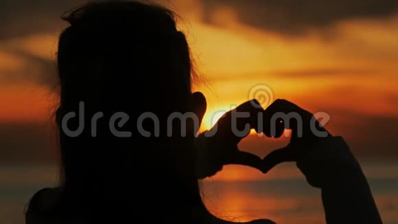 年轻美丽的女孩用她的双手在心形框架下夕阳在海洋上空情感观念视频的预览图