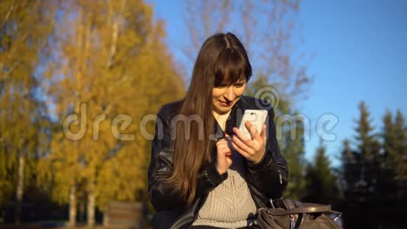 一位年轻的黑发女人在秋天公园里坐在长凳上用电话视频的预览图
