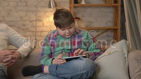 年轻的胖男孩和他的祖父坐在沙发上在平板电脑上玩世代的差异视频的预览图