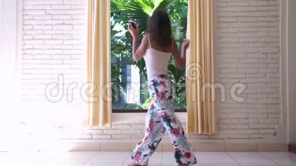 年轻快乐的女人穿着睡衣在窗前跳舞手里拿着手机听着音乐唱着歌享受着音乐视频的预览图