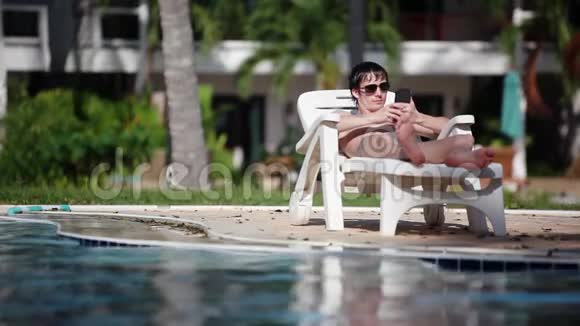 戴墨镜的年轻人在游泳池背景下躺在日光浴床上使用手机1920x1080高清视频的预览图