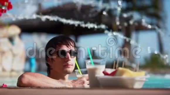 年轻的帅哥戴着太阳镜喝着鸡尾酒吃着热带水果旅行时可以在游泳池放松一下视频的预览图