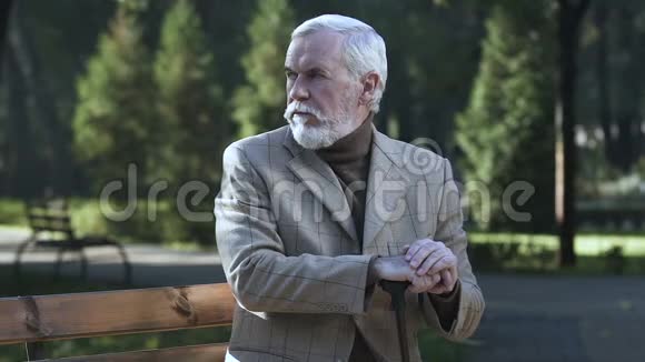 悲伤的养老者坐在公园的长凳上靠在手杖上晚年孤独视频的预览图