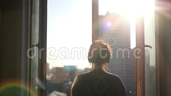 一个年轻的女孩站在敞开的窗户里享受着城市里春天的阳光有镜头的效果慢动作视频的预览图