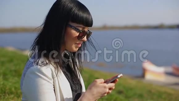 可爱的女士带着太阳镜和长发使用移动设备享受阳光和温暖的白天户外肖像视频的预览图