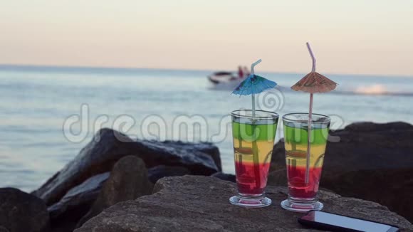 两杯加吸管的鸡尾酒和石头上的智能手机海岸船在后台漂浮视频的预览图