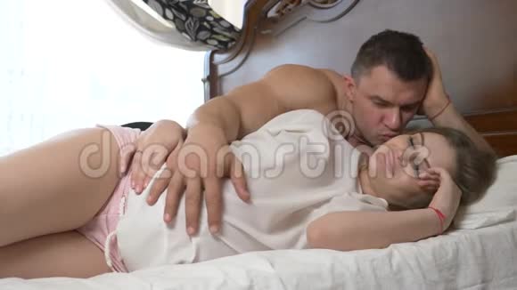 一位年轻漂亮的孕妇和她肌肉发达的丈夫躺在床上丈夫正在熨他妻子的腹部视频的预览图