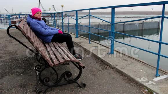 在寒冷的阴天坐在码头上的长凳上的女孩看着镜头视频的预览图