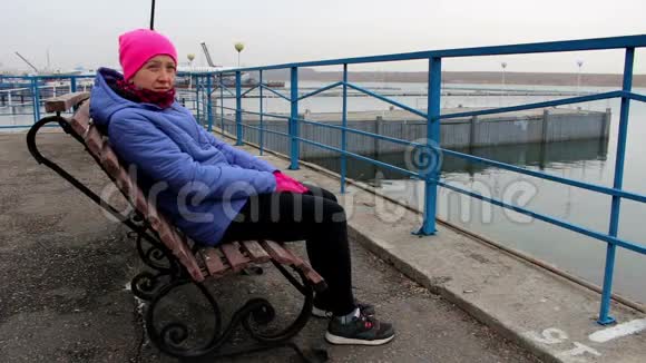 在寒冷的阴天坐在码头上的长凳上的女孩看着镜头视频的预览图